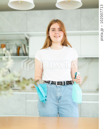 Joyful smiling teen girl holding spray and microfiber rag before cleaning cozy kitchen 102007713