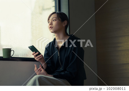 窓辺でスマホを手にコーヒーを飲む物憂げな顔をした若い女性 102008011