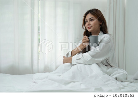 Young woman relaxing and drinking cup of hot coffee or tea in the bedroom Young woman relaxing and drinking cup of hot coffee or tea in the bedroom 102010562