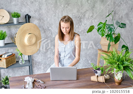 Young female works on a laptop, responds to messages in social networks. Flower delivery concept. Professional worker in a flower shop. Young female works on a laptop, responds to messages in social networks. Flower delivery concept. Professional worker in a flower shop. 102015558