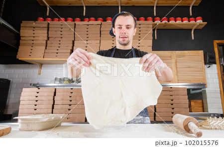 The chef prepares pizza. Raw pizza ready to bake. Cook in a blue apron in the kitchen. with a shovel in his hands. boxes for food delivery on background. 102015576