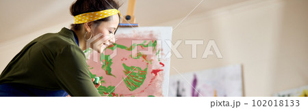 Creative young woman in apron painting a picture on easel, applying paint on canvas with fingers, standing in living room at home studio Creative young woman in apron painting a picture on easel, applying paint on canvas with fingers, standing in living room at home studio 102018133