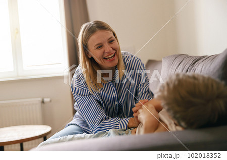 Overjoyed mother having fun with son at home, tickling and playing game on cozy couch in living room 102018352