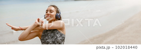 Beautiful young sportive woman in headphone making stretching exercises on beach. Blurred background 102018440