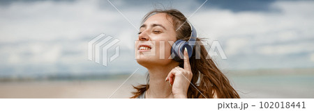 Smiling woman in sportswear listening music on headphones by the beach on background of ocean 102018441