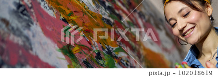 Cheerful young female painter creating a large modern abstract oil painting, applying paint on canvas with fingers in workshop 102018619
