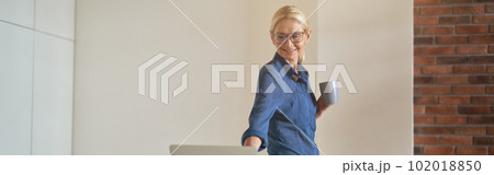 Cheerful mature businesswoman smiling, drinking coffee and using laptop computer while standing in modern kitchen room 102018850