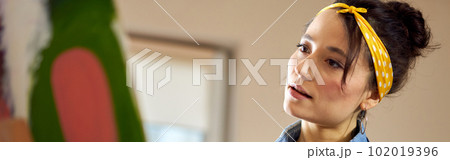 Portrait of young woman in apron looking inspired while working on abstract painting at cozy workshop Portrait of young woman in apron looking inspired while working on abstract painting at cozy workshop 102019396