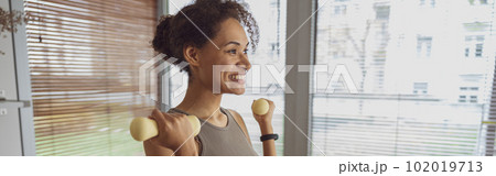 Smiling woman with dumbbells doing exercising at home and working on arms strength 102019713