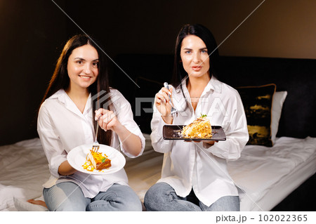 Cheerful girls eating cakes, sitting on bedroom in apartment 102022365
