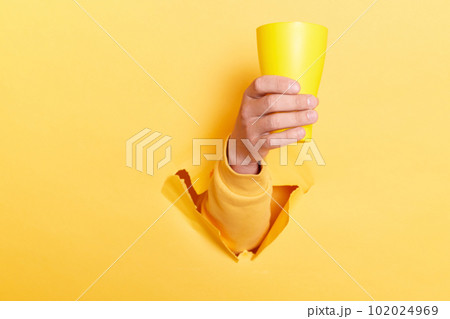 Indoor shot of person hand breaking through yellow paper and holding cup with water or other liquid, showing mug with coffee or tea. 102024969