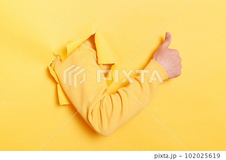 Indoor shot of human hand breaks arm through paper and showing a thumb up sign through a ripped hole, like gesture, isolated over yellow paper background. Indoor shot of human hand breaks arm through paper and showing a thumb up sign through a ripped hole, like gesture, isolated over yellow paper background. 102025619