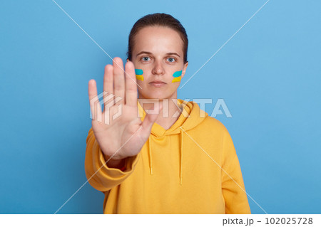 Indoor shot of serious woman in yellow hoodie with Ukrainian flag on cheeks, showing stop gesture with her palm, not war, peace in the world, posing isolated over blue background. Indoor shot of serious woman in yellow hoodie with Ukrainian flag on cheeks, showing stop gesture with her palm, not war, peace in the world, posing isolated over blue background. 102025728
