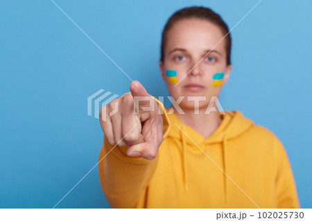 woman wearing yellow hoodie and with flag of Ukraine on her cheek, isolated over blue background, female pointing finger to camera, Stop fighting in Ukraine. Russia stop war. 102025730
