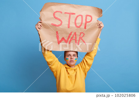 Sad woman holding Ukrainian flag and "Stop the War" placard sign in protest manifestation against Russia invasion on Ukraine. Russian attack, anti-war demonstration. 102025806