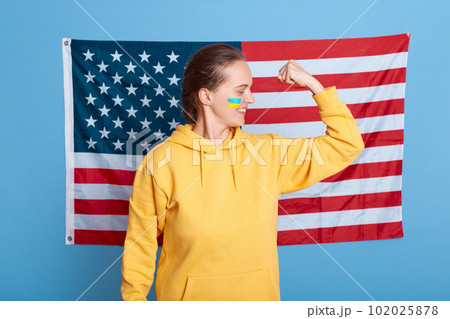 Positive strong woman wearing yellow hoodie and with flag of Ukraine on her cheek, isolated over blue background and american flag, usa support Ukraine, female showing her power, raised her arm. Positive strong woman wearing yellow hoodie and with flag of Ukraine on her cheek, isolated over blue background and american flag, usa support Ukraine, female showing her power, raised her arm. 102025878