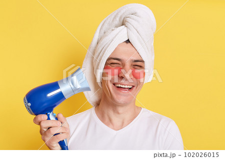 Optimistic happy man wrapped towel on head doing morning cosmetology procedures, drying hair with hair dryer, laughing happily, posing with patches under eyes isolated over yellow background. Optimistic happy man wrapped towel on head doing morning cosmetology procedures, drying hair with hair dryer, laughing happily, posing with patches under eyes isolated over yellow background. 102026015