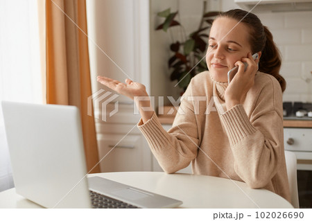 Indoor shot of puzzled woman wearing beige casual style sweater with ponytail hairstyle sitting at table in front of laptop and talking phone, shrugging and doesn't know what to answer. Indoor shot of puzzled woman wearing beige casual style sweater with ponytail hairstyle sitting at table in front of laptop and talking phone, shrugging and doesn't know what to answer. 102026690