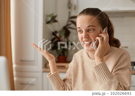 Portrait of angry crazy young adult woman wearing beige sweater sitting in kitchen and talking via mobile phone, spreading hand aside and screaming with hate. 102026691