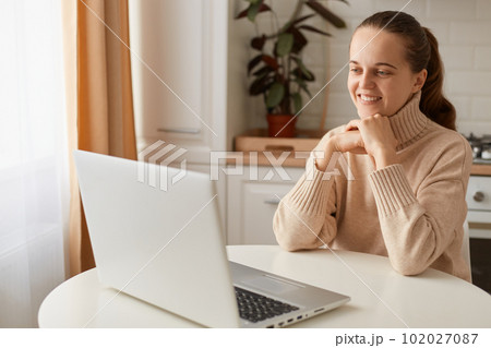 Indoor shot of smiling woman wearing beige sweater sitting in kitchen and having online lesson on laptop, looking at notebook screen with pleasant smile, having video call. 102027087