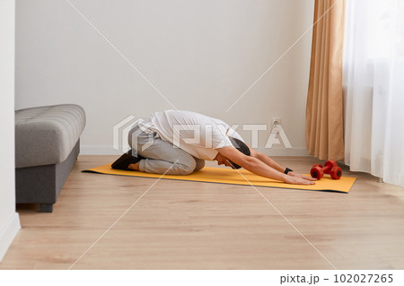 Image of sporty man stretching and practicing yoga at home, young man doing exercises, posing indoor at home on yoga mat, training flexibility, wearing white t shirt and gray pants. 102027265