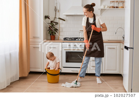 Full length portrait of young adult housewife cleaning house with her little cute charming daughter sitting in bucket, having fun with child while washing floor in kitchen. 102027507