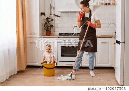 Full length shot of nice attractive hardworking woman making fast domestic work with her cute baby daughter in bucket, wiping floor in modern light white interior kitchen. 102027508