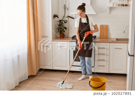 Indoor shot of happy Caucasian woman housewife cleaning floor with mop in kitchen, wearing white t shirt, jeans and brown apron, expressing positive emotions while washing her flat. Indoor shot of happy Caucasian woman housewife cleaning floor with mop in kitchen, wearing white t shirt, jeans and brown apron, expressing positive emotions while washing her flat. 102027891