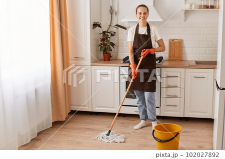Full length portrait of attractive cheerful hardworking woman making fast domestic work, washing floor in modern light white interior kitchen, wearing brown apron, looking at camera. Full length portrait of attractive cheerful hardworking woman making fast domestic work, washing floor in modern light white interior kitchen, wearing brown apron, looking at camera. 102027892