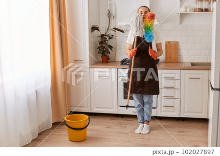 Portrait of young adult Caucasian woman wearing jeans and brown apron, standing in modern kitchen, holding mop and ppduster, washing floor and wiping the dust. 102027897