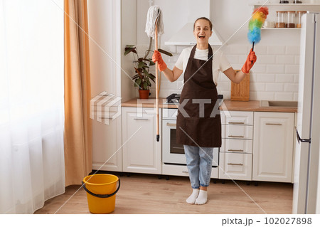 Portrait of excited woman wearing brown apron making fast domestic work wiping and washing floor in modern kitchen, being in good mood, raised arms with her equipment, finishing her cleaning. Portrait of excited woman wearing brown apron making fast domestic work wiping and washing floor in modern kitchen, being in good mood, raised arms with her equipment, finishing her cleaning. 102027898