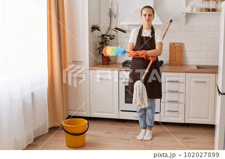 Full length portrait of tired housewife washing floor at kitchen, pretty young woman doing home cleanup, keeping her apartment tidy, feeling overworked, looking at camera. Full length portrait of tired housewife washing floor at kitchen, pretty young woman doing home cleanup, keeping her apartment tidy, feeling overworked, looking at camera. 102027899