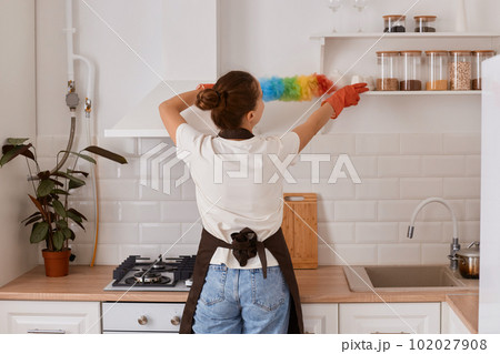 Back view of hardworking woman making domestic work cleansing shelf in kitchen everyday duty in modern light white interior kitchen, wearing white t shirt and jeans. 102027908