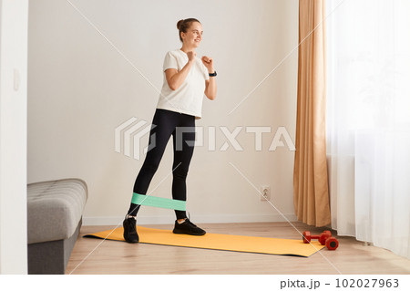 Full and lower body workout. Sporty beautiful woman exercising with elastic rubber band lifting and kicking leg aside standing on yoga mat, Bodyweight training 102027963