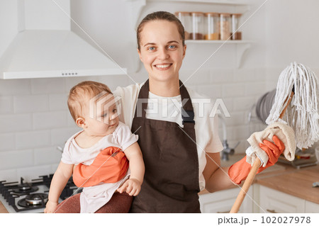 Smiling happy woman with dark hair wearing white t shirt and brown apron, holding mop in hands, holding her baby daughter, housekeeper doing her work child, expressing positive emotions. Smiling happy woman with dark hair wearing white t shirt and brown apron, holding mop in hands, holding her baby daughter, housekeeper doing her work child, expressing positive emotions. 102027978