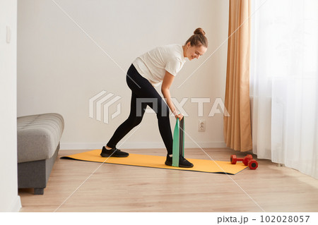 Portrait of sporty woman at home trying to lose weight, standing on rubber stripe training legs, exercising with rubber expander indoor, resistance band for muscle stretching and strength workouts. Portrait of sporty woman at home trying to lose weight, standing on rubber stripe training legs, exercising with rubber expander indoor, resistance band for muscle stretching and strength workouts. 102028057