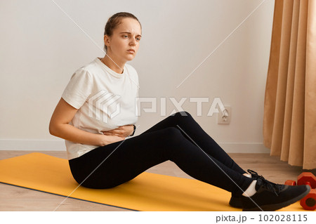 Full length side view portrait of unhealthy sick woman wearing white t shirt and black leggins doing sport exercises at home, sitting on yoga mat and touching painful belly. Full length side view portrait of unhealthy sick woman wearing white t shirt and black leggins doing sport exercises at home, sitting on yoga mat and touching painful belly. 102028115