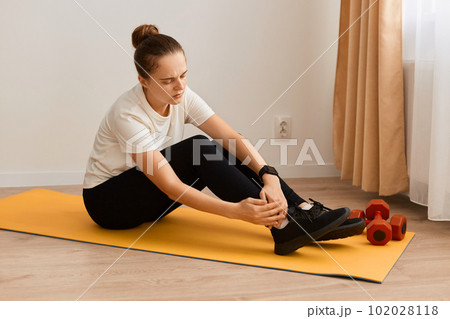 Portrait of ill sick athletic woman wearing white t shirt and black leggins doing sport exercises at home, sitting on mat and keeping hand on painful leg, trauma on workout. 102028118