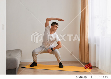 Strong man wearing sportswear and sneakers standing squatting on mat practicing yoga, bending to side with raised hand, doing stretching workouts, looking aside with concentrated expression. Strong man wearing sportswear and sneakers standing squatting on mat practicing yoga, bending to side with raised hand, doing stretching workouts, looking aside with concentrated expression. 102028339