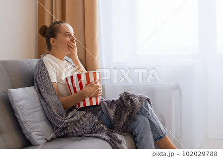 Side view portrait of young adult woman spending time at home sitting on sofa in living room, watching television, scary or horror movie, in panic eating popcorn bowl. Side view portrait of young adult woman spending time at home sitting on sofa in living room, watching television, scary or horror movie, in panic eating popcorn bowl. 102029788