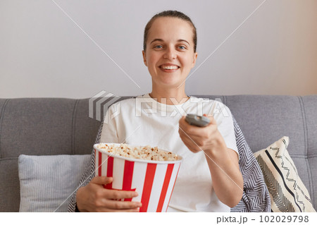 Image of happy satisfied woman wearing white casual style t shirt, sitting on sofa and eating popcorn, watching comedian film alone, smiling happily at camera. Image of happy satisfied woman wearing white casual style t shirt, sitting on sofa and eating popcorn, watching comedian film alone, smiling happily at camera. 102029798