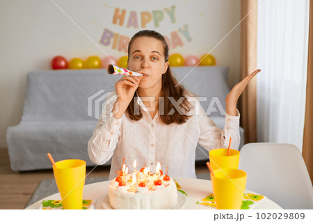 Portrait of happy Caucasian birthday lady blowing party tube while sitting at table in decorated living room, expressing positive emotions, looking at camera and spreading hand. Portrait of happy Caucasian birthday lady blowing party tube while sitting at table in decorated living room, expressing positive emotions, looking at camera and spreading hand. 102029809