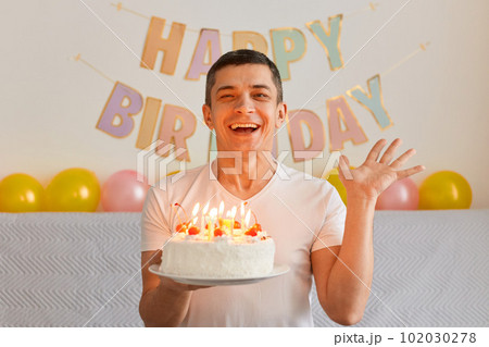 Indoor shot of brunette joyful young adult man holding birthday cake and looking at the camera at home, waving at camera with excitement, posing at home with festive interior decoration on background. 102030278