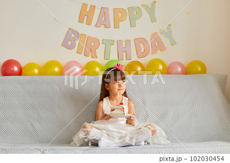 Indoor shot of adorable female child wearing white dress sitting on cough with balloons and festive inscriptions on background, holding cake, looking away and making wish, celebrating birthday. 102030454