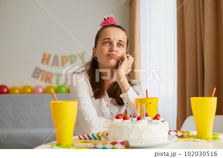 Image of bored attractive young woman wearing white shirt celebrating birthday, sitting alone at table with cake, keeping hand under chin, looking away, dreaming. Image of bored attractive young woman wearing white shirt celebrating birthday, sitting alone at table with cake, keeping hand under chin, looking away, dreaming. 102030516