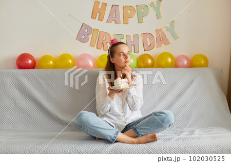 Pensive woman wearing white shirt and jeans sitting on sofa with crossed legs, holding cake with candle, looking aside and making wish, celebrating birthday, festive decoration on background. 102030525