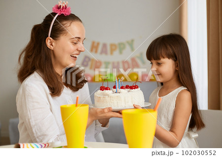 Indoor shot of woman with daughter cerebrating birthday, mother holding cake and congratulating little girl, family posing at home with festive inscription and balloons on background. Indoor shot of woman with daughter cerebrating birthday, mother holding cake and congratulating little girl, family posing at home with festive inscription and balloons on background. 102030552