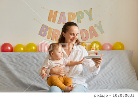 Horizontal shot of happy smiling woman sitting on sofa with her infant daughter and holding cake with candles, smiling happily, expressing positive emotions, celebrating first baby birthday. Horizontal shot of happy smiling woman sitting on sofa with her infant daughter and holding cake with candles, smiling happily, expressing positive emotions, celebrating first baby birthday. 102030633