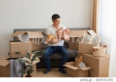 Portrait of Caucasian man wearing white T-shirt and jeans sitting on sofa with toddler daughter surrounded with cardboard boxes, calming down crying baby kid, moving in a new home with family. Portrait of Caucasian man wearing white T-shirt and jeans sitting on sofa with toddler daughter surrounded with cardboard boxes, calming down crying baby kid, moving in a new home with family. 102031185