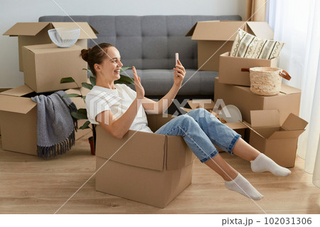 Indoor shot of kind happy woman wearing white t shirt and jeans sitting in a cardboard box and having video call on smart phone, showing hello gesture, waving hand. Indoor shot of kind happy woman wearing white t shirt and jeans sitting in a cardboard box and having video call on smart phone, showing hello gesture, waving hand. 102031306